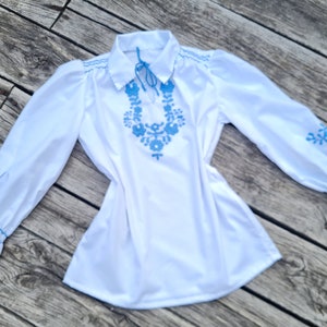 May include: White long-sleeved blouse with blue embroidered floral details around the neckline and on the cuffs.
