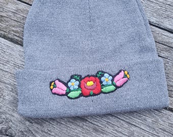 Puede incluir: Un gorro gris con bordado floral. El bordado incluye tulipanes rosas, nomeolvides azules y una rosa roja. El gorro está hecho de un material de punto suave y es adecuado para el clima frío.