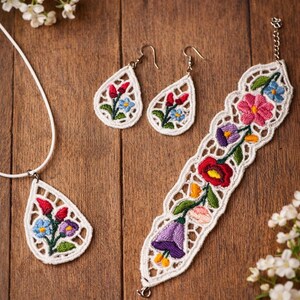May include: A white lace jewellery set with colourful floral embroidery. The set includes a pendant necklace, a pair of teardrop earrings, and a bracelet. The embroidery features red, blue, purple, and yellow flowers. The jewellery is displayed on a wooden surface.