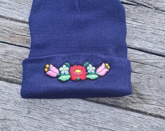 Puede incluir: Un gorro de punto azul marino con un diseño de bordado floral. Las flores bordadas incluyen detalles en rosa, rojo, azul y amarillo. El gorro se muestra sobre una superficie de madera envejecida.