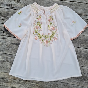 May include: A white sheer blouse with embroidered pink and green floral designs. The blouse has a gathered neckline and short sleeves with crochet trim.