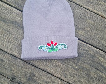 Puede incluir: Un gorro gris claro con un diseño floral bordado en la parte delantera. El bordado presenta flores rojas y detalles azules y verdes. El gorro está hecho de un material suave y tejido.