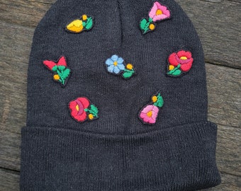 Puede incluir: Un gorro gris oscuro adornado con parches florales bordados en varios colores. Las flores incluyen rosa, rojo, amarillo y azul, añadiendo un toque de color al gorro. El gorro está hecho de un material de punto.