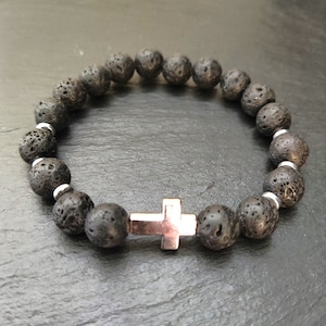 Könnte beinhalten: Ein schwarzes Armband aus Lavasteinen mit einem silbernen Kreuzanhänger. Das Armband besteht aus kleinen, runden Perlen und hat ein schlichtes, elegantes Design.