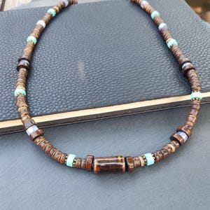 Può includere: Una collana di perline di legno marrone con accenti turchesi e viola. La collana ha una perla cilindrica di legno marrone al centro.