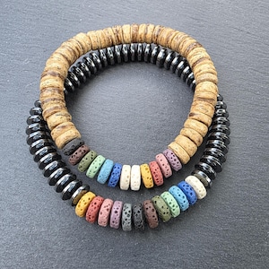 May include: Two stacked bracelets. The top bracelet features light brown wooden beads. The bottom bracelet has black, metallic beads. A second bracelet, with colourful beads, is stacked on top of the black bracelet. The colourful beads include red, green, blue, and yellow.