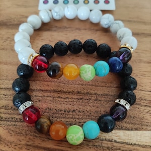 Op de afbeelding: Twee armbanden, een van zwarte lavasteen en een witte, met gekleurde chakra- kralen. De armbanden liggen op elkaar.