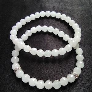 Op de afbeelding: Twee witte kralen armbanden met zilveren accenten. De armbanden zijn gemaakt van ronde, witte kralen en hebben een zilveren band met kleine, heldere stenen op elke armband.