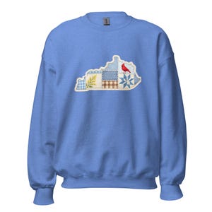 Peut inclure: Un sweat-shirt bleu clair orné d'un motif patchwork en forme du Kentucky. Le patchwork comprend divers motifs et un cardinal rouge. Le sweat-shirt a des manches longues et un col rond.