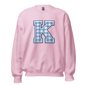 Peut inclure: Un sweat-shirt rose clair avec une grande lettre "K" brodée. La lettre est dans un motif vichy bleu et blanc, bordée d'un liseré bleu plus foncé et de coutures blanches. Le sweat-shirt a un col rond et des manches longues.