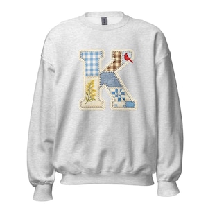May include: A pale grey sweatshirt with a large, patchwork letter "K". The letter is made up of different fabric patterns, including gingham, floral, and denim. A small red bird sits on the upper right of the letter. The sweatshirt has ribbed cuffs and a crew neck.
