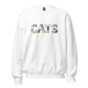 Peut inclure: Sweat-shirt blanc avec le mot "CATS" en patchwork. Les lettres sont faites de tissu bleu et blanc à motifs, avec un bord jaune. Le sweat-shirt a un col rond et des manches longues.