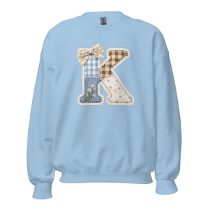 Op de afbeelding: Een lichtblauwe sweatshirt met een grote patchwork letter "K". De letter is versierd met een ruitjesstrik en diverse stofpatronen in blauw, bruin en wit, met bloemen- en stippeldetails. Een comfortabele, casual stijl.