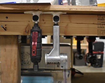 Può includere: Due utensili elettrici sono appesi a un banco da lavoro in legno. Gli utensili sono montati su supporti in plastica bianca con cuscinetti in gomma nera. L'utensile a sinistra è una smerigliatrice angolare rossa e nera. L'utensile a destra è una levigatrice a nastro argentata.