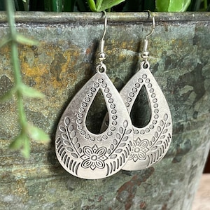 May include: A pair of silver-colored teardrop earrings. Each earring features a floral design with a central flower and leaf patterns. The edges are adorned with small circular details, and the earrings hang from silver hooks.