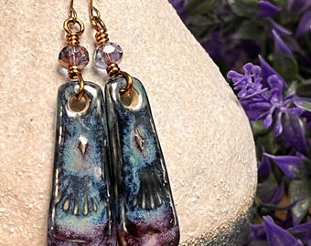 May include: Artisan-made earrings with a unique design. These dangle earrings feature rectangular ceramic pendants with a blend of blue, purple, and black glaze. The earrings are accented with copper wire and small, faceted beads. A stylish accessory.