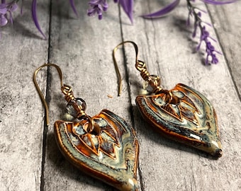 May include: A pair of handcrafted earrings with a leaf-like design. The earrings feature a blend of brown, blue, and cream colors. The ear wires are gold-colored, and the earrings are displayed against a wooden background with purple flowers.