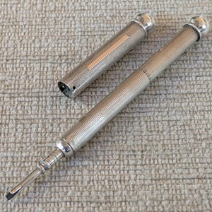May include: A silver-toned mechanical pencil with a fine vertical ribbed texture along the barrel and cap. The pencil features a rounded top, a smooth metallic band for engraving, and a slender, pointed tip extending from the main body.