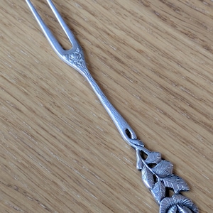 May include: A silver-toned metal cocktail fork featuring a two-pronged head and a long, slender handle. The handle is decorated with a detailed, raised rose flower motif at the base and a smaller rose design where the handle meets the fork tines.