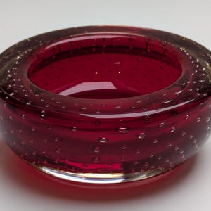 May include: A circular, deep red glass bowl with a thick rim and a clear base. The bowl is filled with tiny air bubbles throughout the glass. A decorative item.