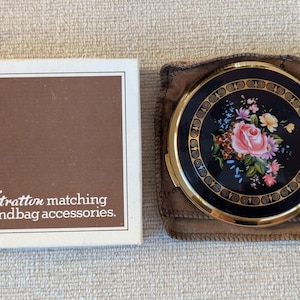 May include: A round gold-tone compact mirror with a black enamel lid featuring a floral bouquet of pink roses and blue flowers. The compact is housed in a brown fabric pouch next to a white box labelled Stratton matching handbag accessories.