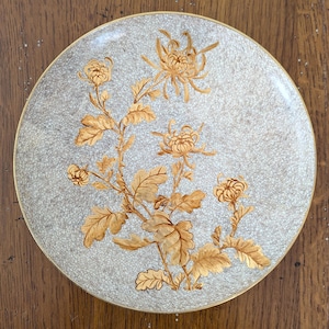 Puede incluir: Un plato redondo de cerámica con fondo beige claro y un patrón de esmalte craquelado oscuro y fino. El centro está decorado con una ilustración detallada de flores y hojas de crisantemo de color marrón dorado, con un borde fino dorado.