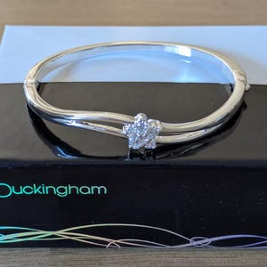 Peut inclure: Un bracelet jonc articulé de couleur argentée orné d'un motif central en forme de fleur incrusté de pierres transparentes. Le bracelet repose sur une boîte rectangulaire noire avec le mot buckingham imprimé en bleu clair et un motif linéaire coloré.
