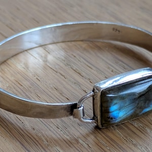 May include: A silver bangle bracelet featuring a rectangular labradorite gemstone set in a polished metal frame. The stone displays a prominent iridescent blue flash against a dark grey base. The band is smooth, solid, and metallic.