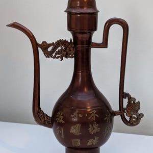 May include: Bronze-coloured decorative pitcher with a long spout and a curved handle. The pitcher features ornate dragon and floral embellishments, along with a small lion figure on the lid. The body of the pitcher has engraved characters.
