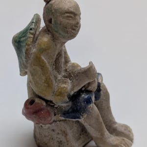 Op de afbeelding: Een kleine, verouderde figuur van een zittende figuur met een hoed en vleugels. De figuur is beschilderd met gedempte kleuren, waaronder blauw, geel en rood. De figuur lijkt gemaakt van keramiek of een vergelijkbaar materiaal.
