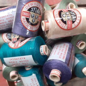 May include: A collection of spools of thread in various colours, including teal, purple, beige, and green. The spools are labelled with the brand name "Sylko" and the text "Three Machine Twist Shells".