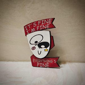 May include: A wooden cutout of a face with the text "It's fine I'm fine Everything's fine" in a red banner. The face has a black eye, a spiral, a star, and a smile.