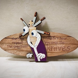 May include: A wooden oval plaque featuring a cartoon chess king wearing a purple robe with white trim, large googly eyes, and knives protruding from its head. The text reads: Can you repeat the question? I couldn't hear you over the knives.