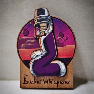 May include: Wooden art piece depicting a cartoon figure with a bucket-shaped head and purple body, set against a sunset-coloured landscape with planets. The text "the Bucket Whisperer" is carved into the wooden base.