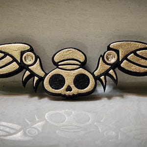 Pode incluir: Um emblema decorativo com uma caveira com grandes asas estilizadas. A caveira e as asas são delineadas em preto com um preenchimento texturizado dourado. O emblema tem um design simétrico e é provavelmente um patch ou aplique.