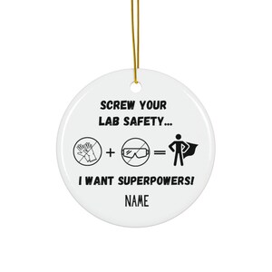 May include: A white ceramic round Christmas ornament with a gold hanging string. The ornament has black text and graphics, including the phrase "SCREW YOUR LAB SAFETY..." and "I WANT SUPERPOWERS!" with a superhero graphic.