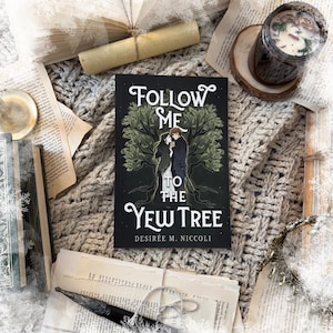 May include: A book titled "Follow Me to the Yew Tree" with a cover illustration of a couple under a tree. The image includes a candle, rolled paper, and scattered book pages, all arranged on a textured grey surface.