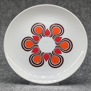 Könnte beinhalten: Ein weißer Keramikteller mit einem braunen und orangefarbenen geometrischen Muster. Das Muster ist eine stilisierte Blume mit acht Blütenblättern.