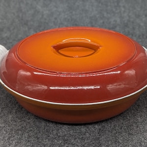 Könnte beinhalten: Orangefarbene und rote emaillierte gusseiserne Auflaufform mit weißen Griffen und einem Deckel. Der Deckel hat einen kleinen orangenen Griff.
