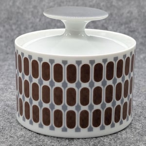 Könnte beinhalten: Eine weiße Zuckerdose aus Keramik mit Deckel. Die Schüssel hat ein sich wiederholendes Muster aus braunen Ovalen und grauen Rechtecken. Der Deckel hat einen kleinen, runden Griff. Es ist ein Küchenartikel.