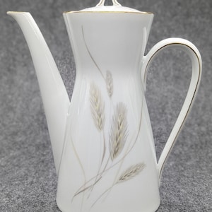 Könnte beinhalten: Weiße Porzellan-Kaffeekanne mit Weizenähren-Design. Die Kanne hat einen langen Ausguss, einen gebogenen Griff und einen Deckel mit Goldrand. Die Weizenähren sind in Beige- und Grautönen gehalten.
