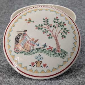 Könnte beinhalten: Runder, weißer Keramikdeckel mit dekorativer Illustration. Der Deckel hat einen roten Rand, ein gelb-blaues Zickzackmuster und eine zentrale Szene mit einem Baum, Figuren und Vögeln. Das Design erinnert an Vintage-Geschirr.
