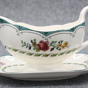 Könnte beinhalten: Eine weiße Porzellan-Soßenkännchen mit grünem und floralem Design. Die Kanne hat einen Griff und steht auf einer passenden Untertasse.