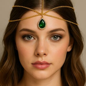 May include: A gold-toned headpiece with a central teardrop-shaped green gemstone. The headpiece features delicate chain details that extend across the forehead and around the head, creating an elegant and eye-catching accessory.