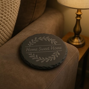 May include: A round, dark gray slate coaster with the phrase "Home Sweet Home" engraved in a decorative font. The coaster features a leaf design around the text. The coaster is sitting on a brown upholstered surface.