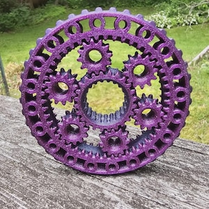 Puede incluir: Un mecanismo de engranajes púrpura impreso en 3D con múltiples engranajes entrelazados de varios tamaños. El anillo exterior presenta recortes circulares, y los engranajes están dispuestos concéntricamente. El objeto está colocado sobre una superficie de madera envejecida.