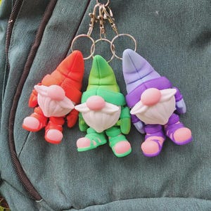 May include: Three colorful gnome keychains. Each gnome has a different color scheme: red, green, and purple. They have white beards, pink noses, and matching shoes and hats. The keychains are attached to a metal ring.