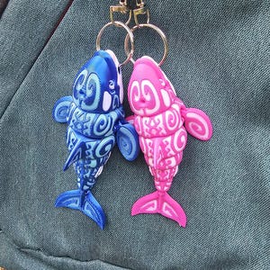 Pode incluir: Dois chaveiros em forma de peixe, um azul e outro rosa, com detalhes brancos intrincados. Cada chaveiro é preso a um chaveiro e fecho prateado. Os peixes têm um design estilizado com barbatanas e caudas. O fundo é um tecido verde texturizado.
