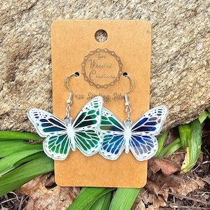 May include: A pair of butterfly earrings with iridescent green, blue, and white wings. The earrings are made of 925 sterling silver and are attached to a card with the text "Two Wheeled Creations".
