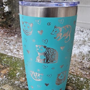 May include: A turquoise stainless steel tumbler with a silver design of opossums and hearts.